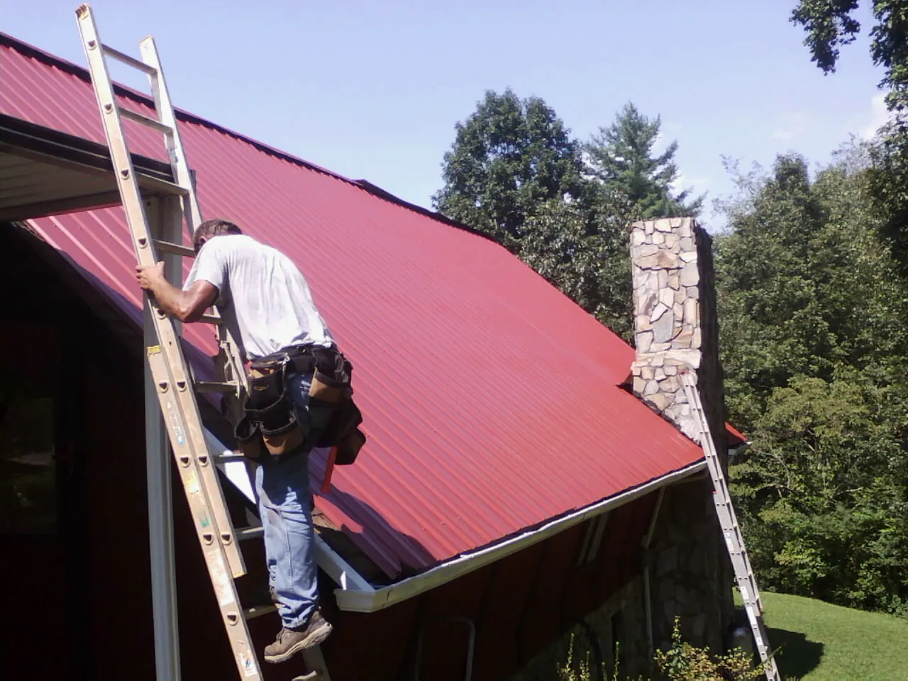 Roofer on a ladder beside a red metal roof and stone chimney — Soffit & Fascia Repair by Roofing Brothers in Wilton